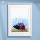 Framed artwork of a colourful hermit crab on sand with a blue watercolor background, displayed on a light blue wall. Artist - Naomi Jenkin. 