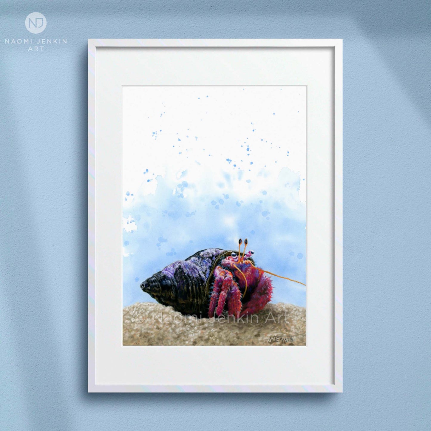 Framed artwork of a colourful hermit crab on sand with a blue watercolor background, displayed on a light blue wall. Artist - Naomi Jenkin. 
