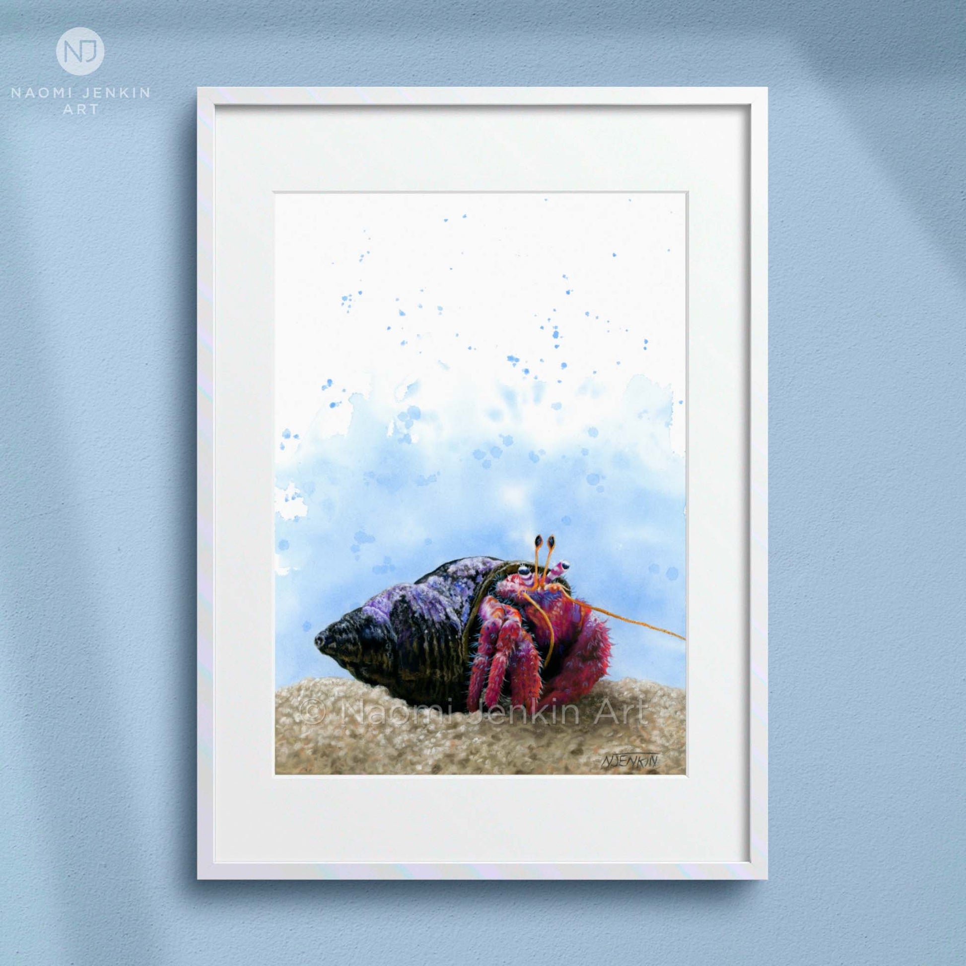 Framed artwork of a colourful hermit crab on sand with a blue watercolor background, displayed on a light blue wall. Artist - Naomi Jenkin. 
