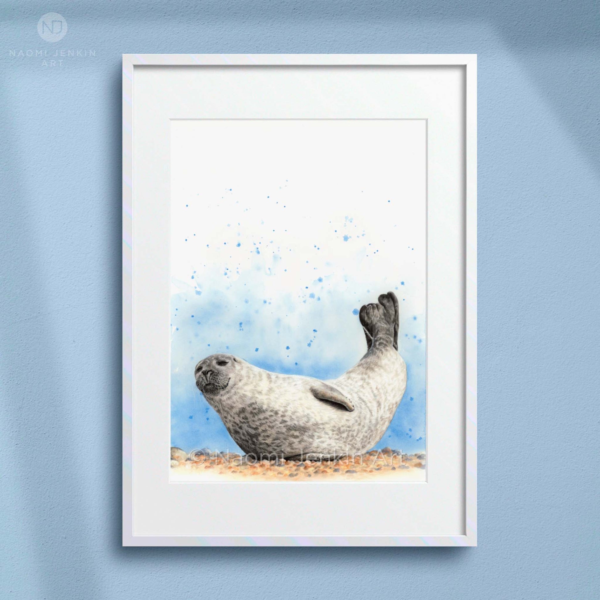 Framed artwork of a seal relaxing on the beach with a blue painted background. Artist - Naomi Jenkin. 
